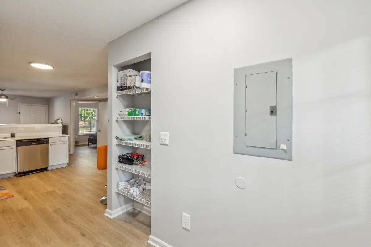 A modern, open kitchen with visible shelving neatly arranged with everyday items. The kitchen flows seamlessly into a living area through a wide doorway, suggesting easy access between spaces. The decor is inviting and thoughtfully arranged, balancing function and style.