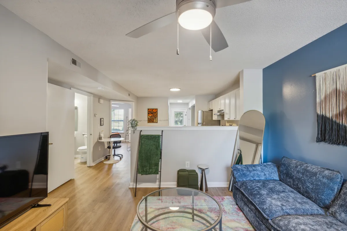 A cozy and inviting living area featuring a plush sofa, coffee table, and adjacent kitchen space. Large windows fill the room with natural light, and tasteful artwork completes the setting, making it ideal for relaxing or entertaining guests.