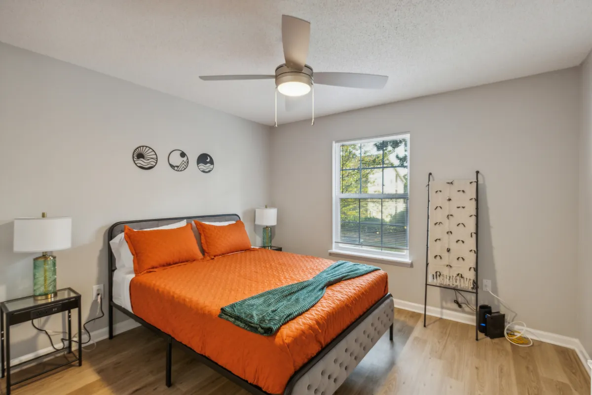 A bright, well-lit bedroom featuring a queen-sized bed with a warm orange quilt and a green throw blanket. Natural light pours through the window, illuminating decorative wall art and a nightstand with a lamp—creating a cozy, restful atmosphere.