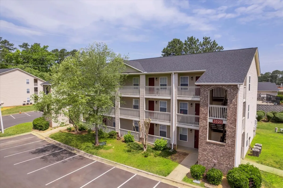A three-story apartment building surrounded by lush green landscaping, with a parking lot in the foreground. The scene suggests a peaceful, well-maintained environment, with balconies hinting at private relaxation spaces and communal gardens.