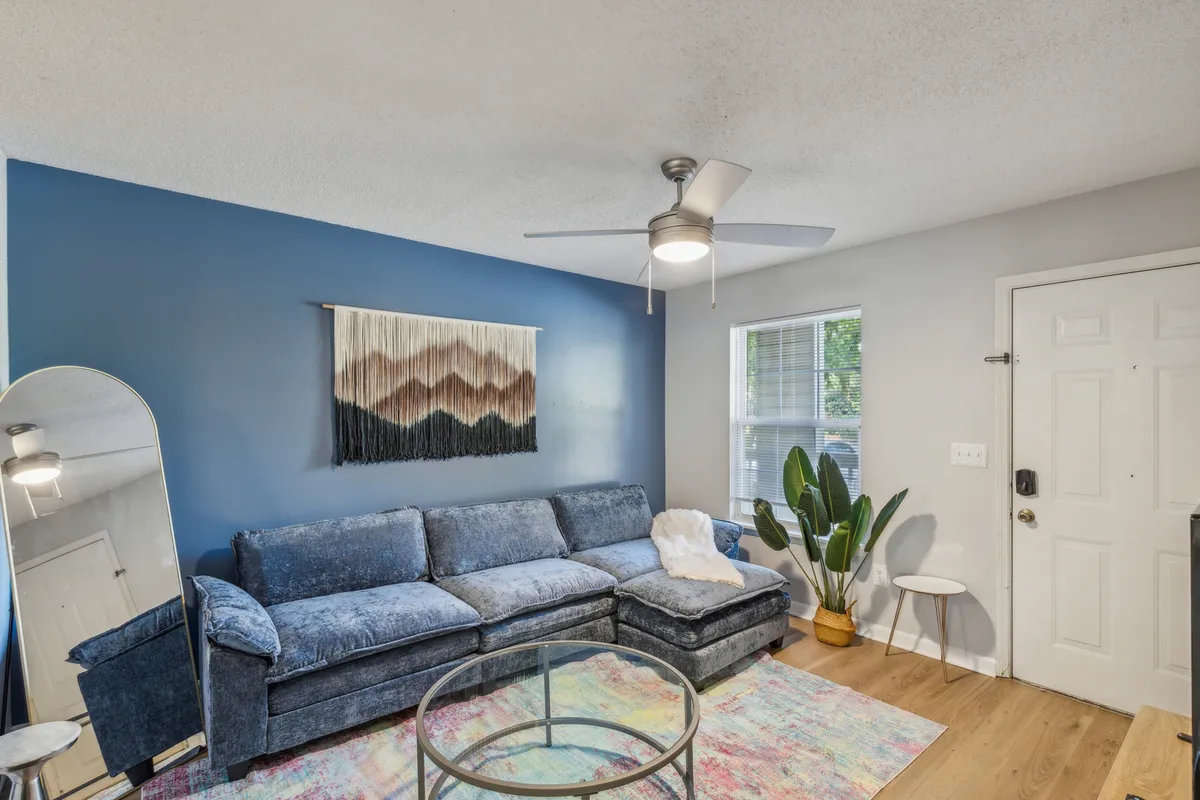 A modern living area with a blue sectional sofa, textured wall hanging, round glass coffee table, and lush indoor plants. The space feels airy and welcoming, designed for both relaxation and socializing.