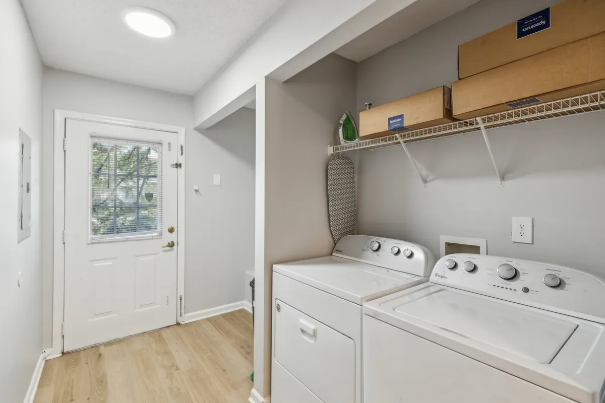 A bright, practical laundry area with a front-loading washing machine and dryer stacked side by side. Organized shelves overhead hold boxes and cleaning supplies, and a door leads to the outside, allowing for natural light and easy movement. The space is tidy and functional, clearly designed for everyday convenience.