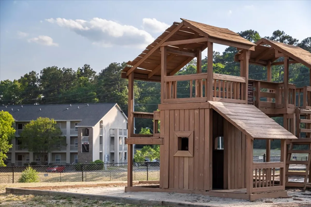 A cheerful exterior scene highlights a playhouse structure surrounded by trees and an apartment building. Children are playing nearby, and families enjoy the open green space, suggesting a strong sense of community and safe, family-oriented environment.