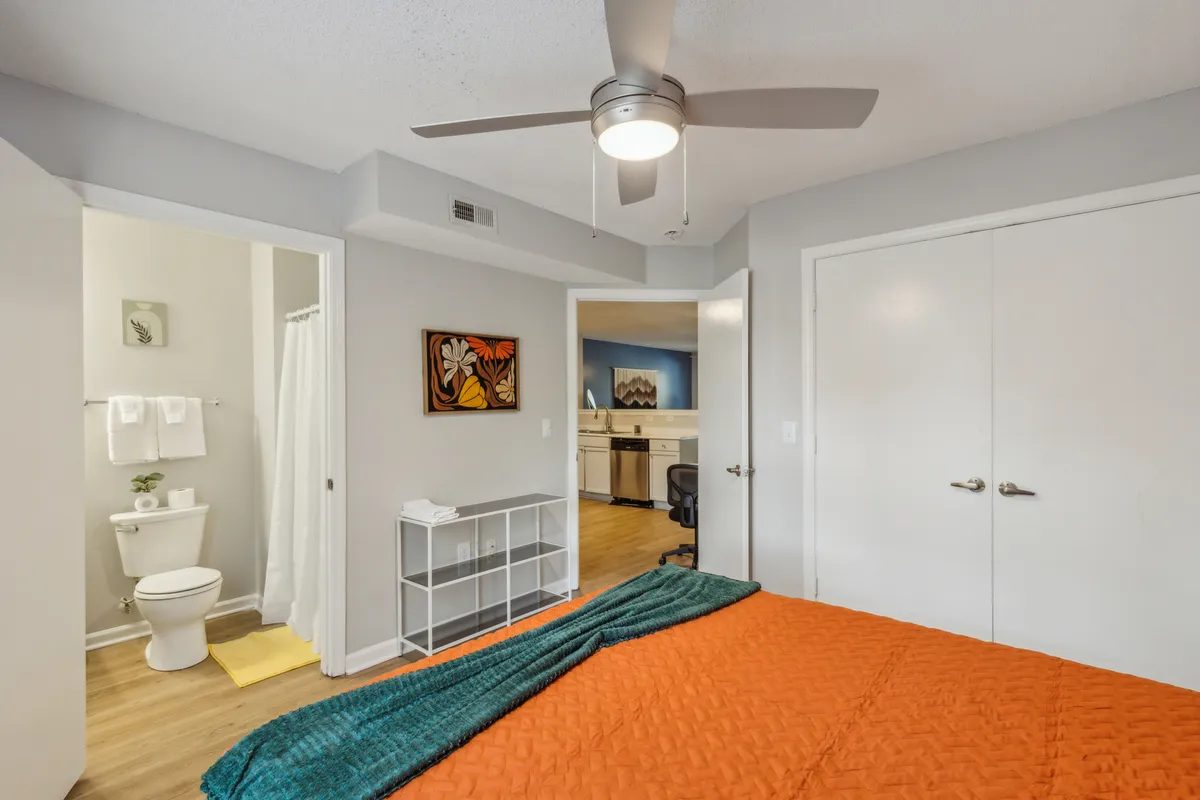 A spacious queen bedroom with an orange quilt, artistic wall décor, a ceiling fan, and a doorway that leads directly into a bright bathroom. The room feels welcoming and airy, with hints of the en-suite visible.