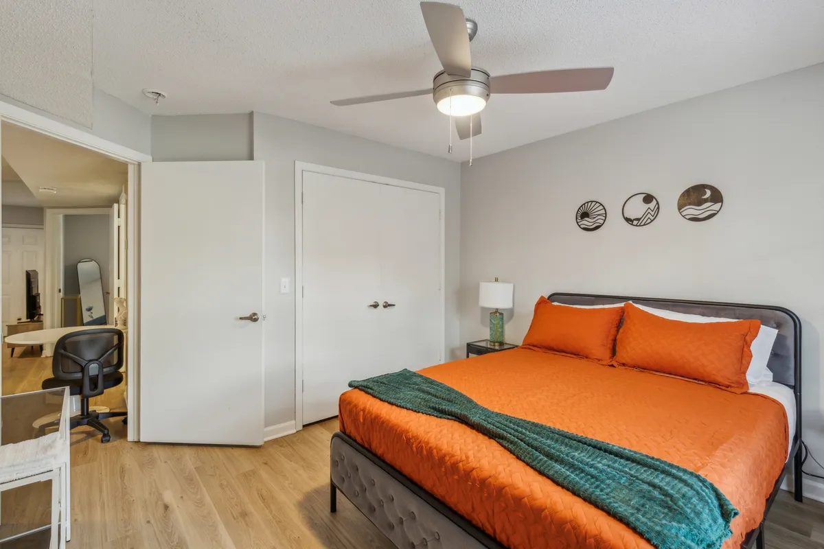A cozy bedroom featuring an orange bedspread, decorative wall art, and a nightstand with a warm lamp. Sunlight streams in, emphasizing the inviting and restful atmosphere. The decor is stylish yet practical, with visible personal touches and storage options.