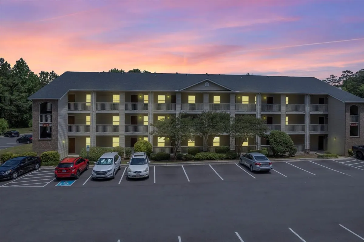 A sunset sky above the building's facade, with parked cars and landscaping visible. The balconies hint at private outdoor space, while the overall mood is warm and welcoming, suggesting a pleasant return after a busy day.